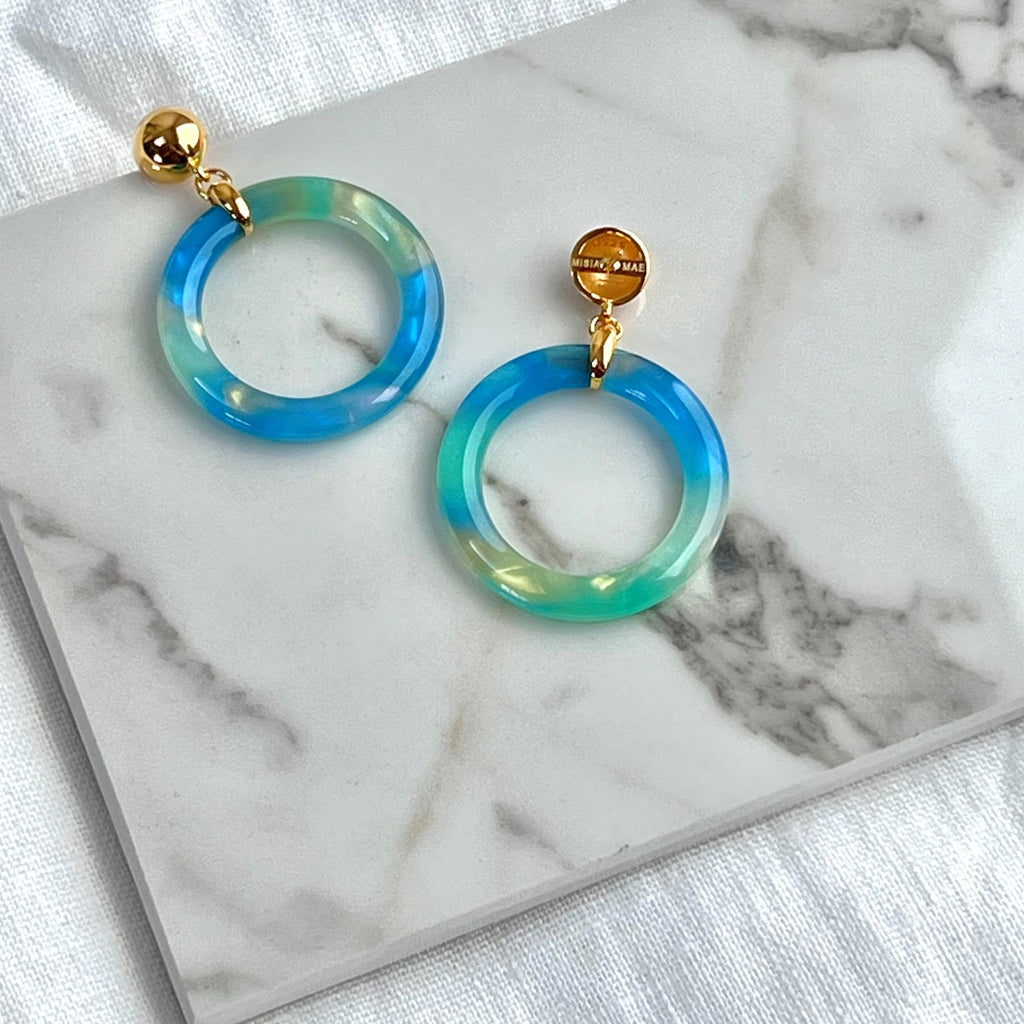 Blue earrings from the Audrey collection  are presented on a white marble top and white linen cloth. The 19502 style earrings are made of tortoiseshell and 14k gold vermeil.