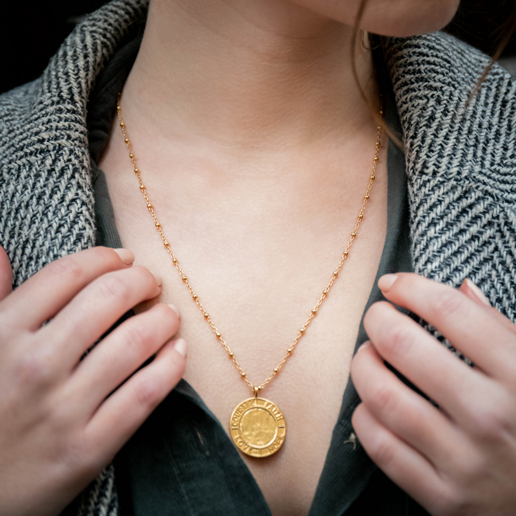 Person wearing a gold necklace with a coin pendant, hands adjusting the garment.